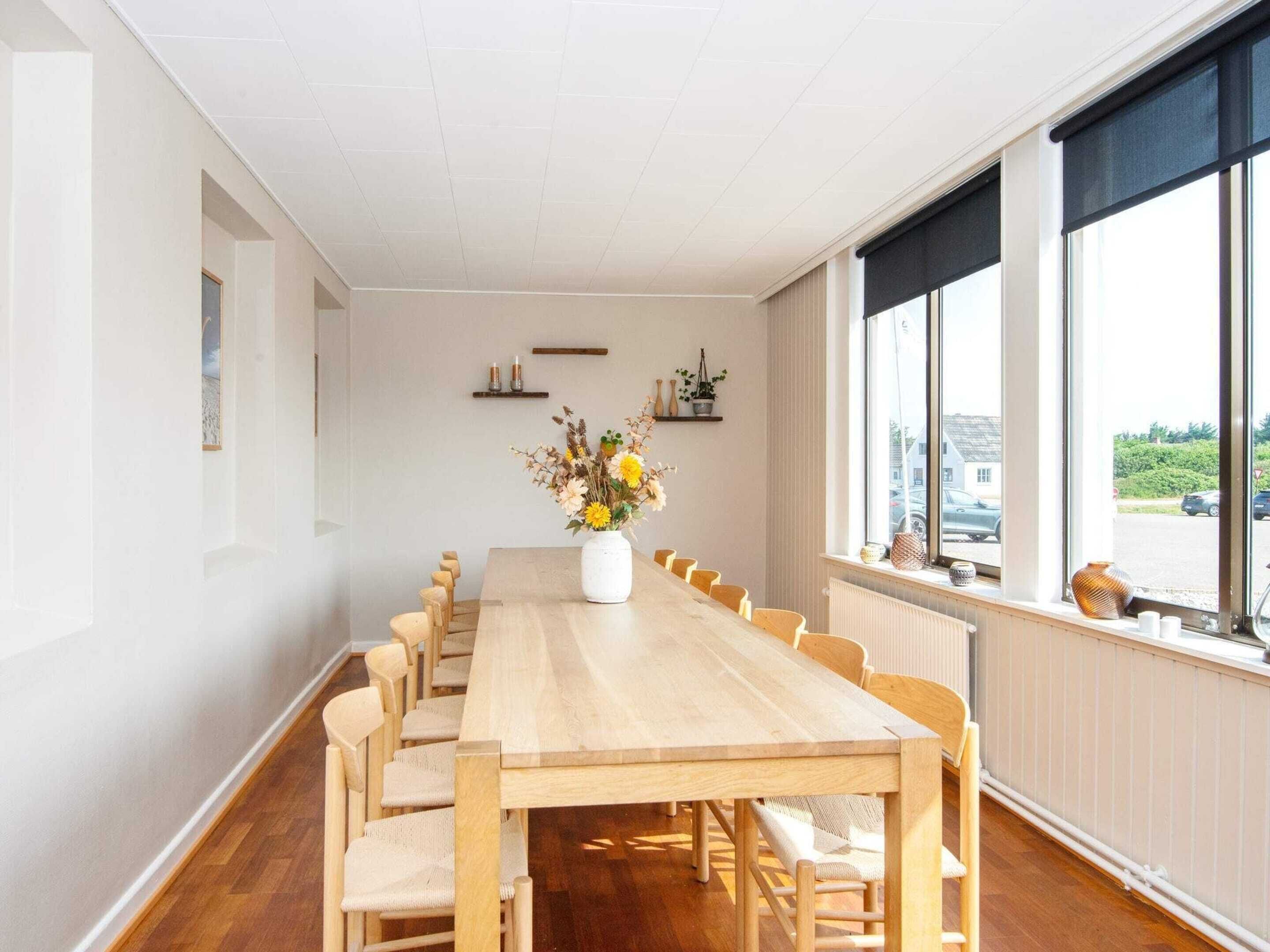 A long dining table with chairs, a vase of flowers, and a view of the street through large windows.