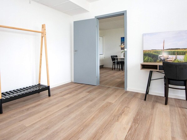 Una habitación con piso de madera, un escritorio con una silla, un cuadro en la pared y una puerta que da a otra habitación.