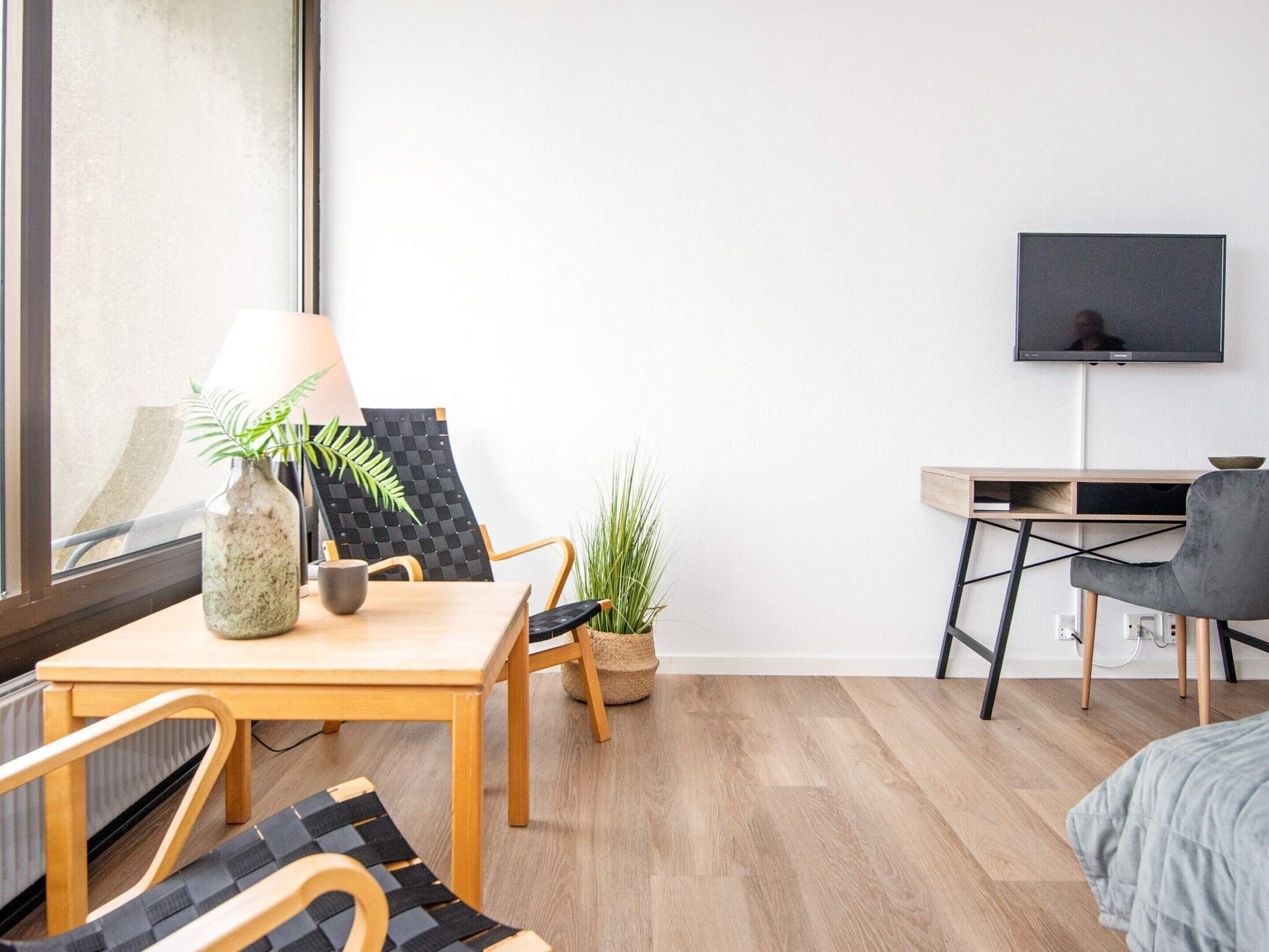 Una habitación moderna y luminosa con una mesa de madera, dos sillas, un escritorio pequeño con una computadora y un televisor, además de una planta en maceta.