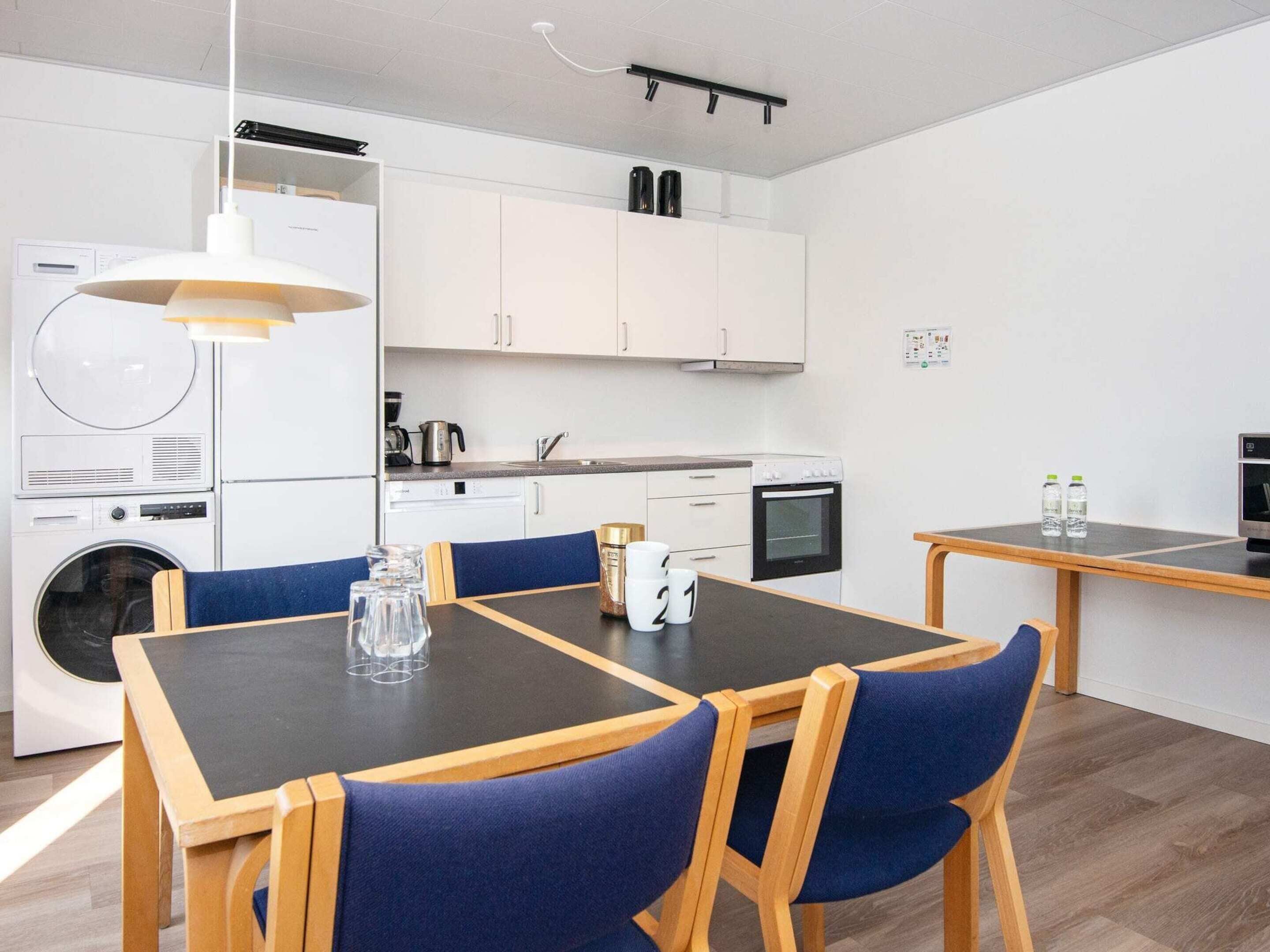 A compact kitchen with white cabinets, a built-in oven, and a coffee machine. There is a dining table with blue chairs and a small table with two bottles on it.