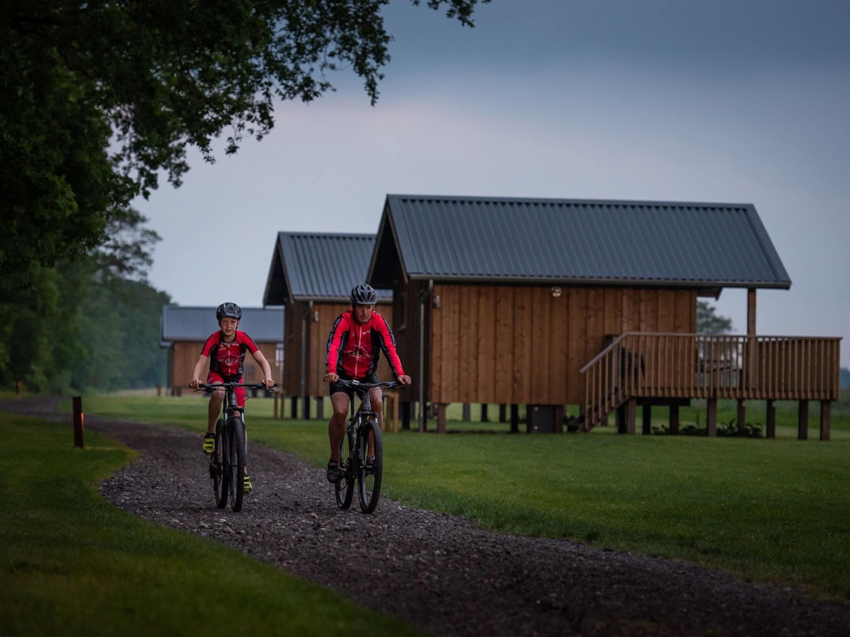 Two cyclists on a gravel path with wooden buildings in the background.