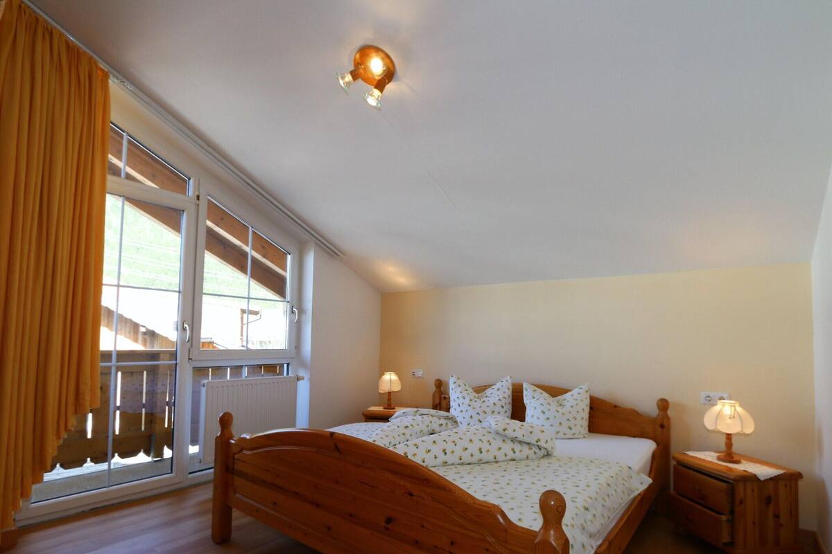 A bedroom with a wooden bed, two bedside tables with lamps, a window with curtains, and a ceiling light fixture.