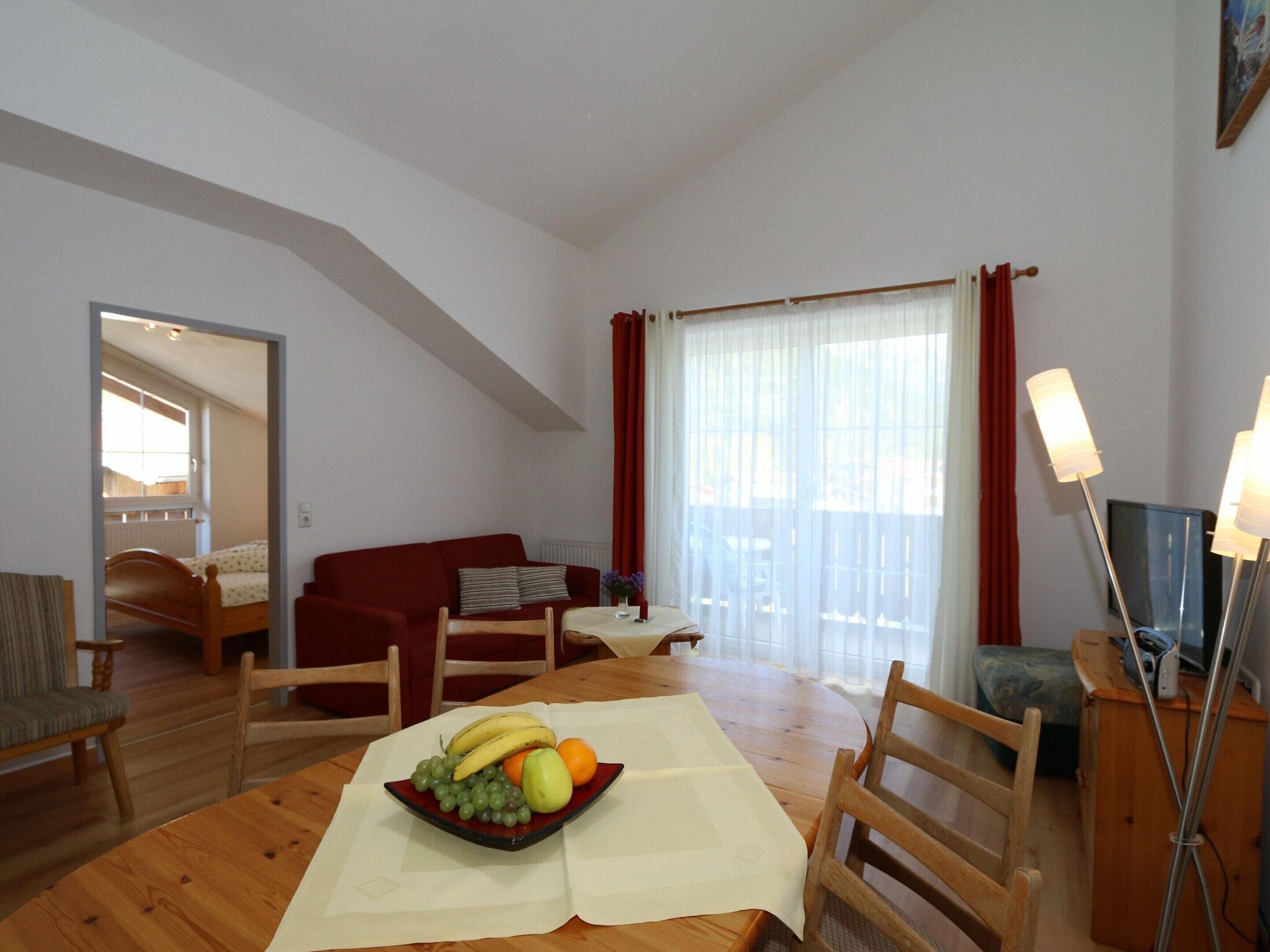 A living room with a wooden dining table, red sofa, and a view of a balcony through a large window with curtains.