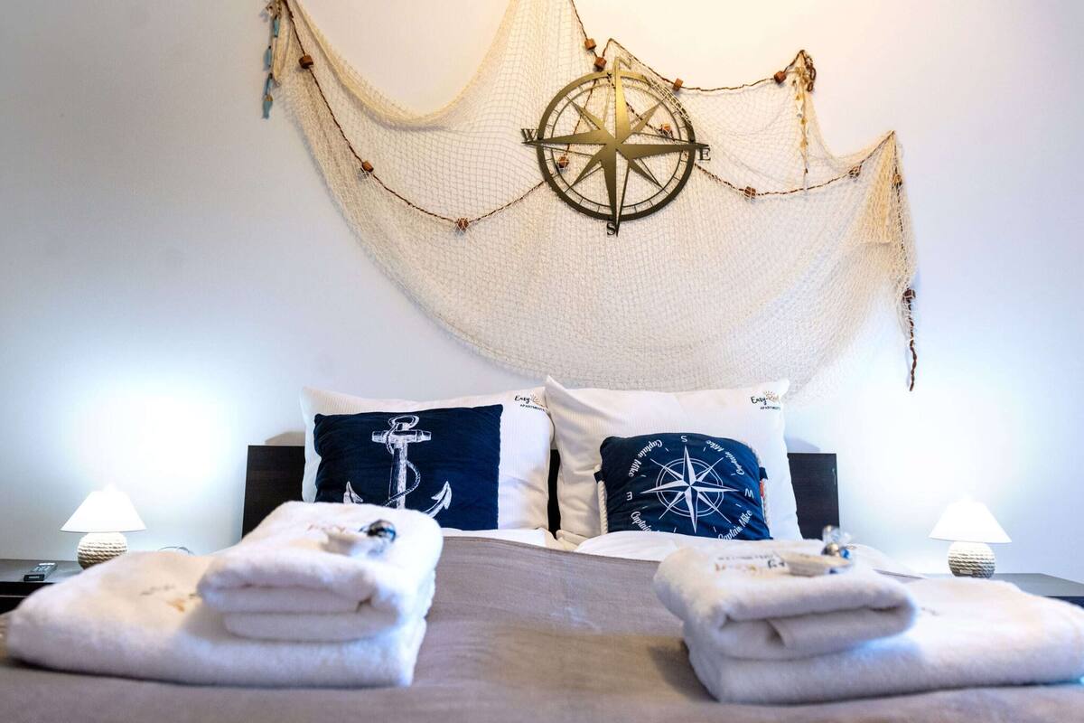 Uma cama bem arrumada com duas almofadas, um compasso decorativo fixado na parede e duas luminárias de cabeceira.