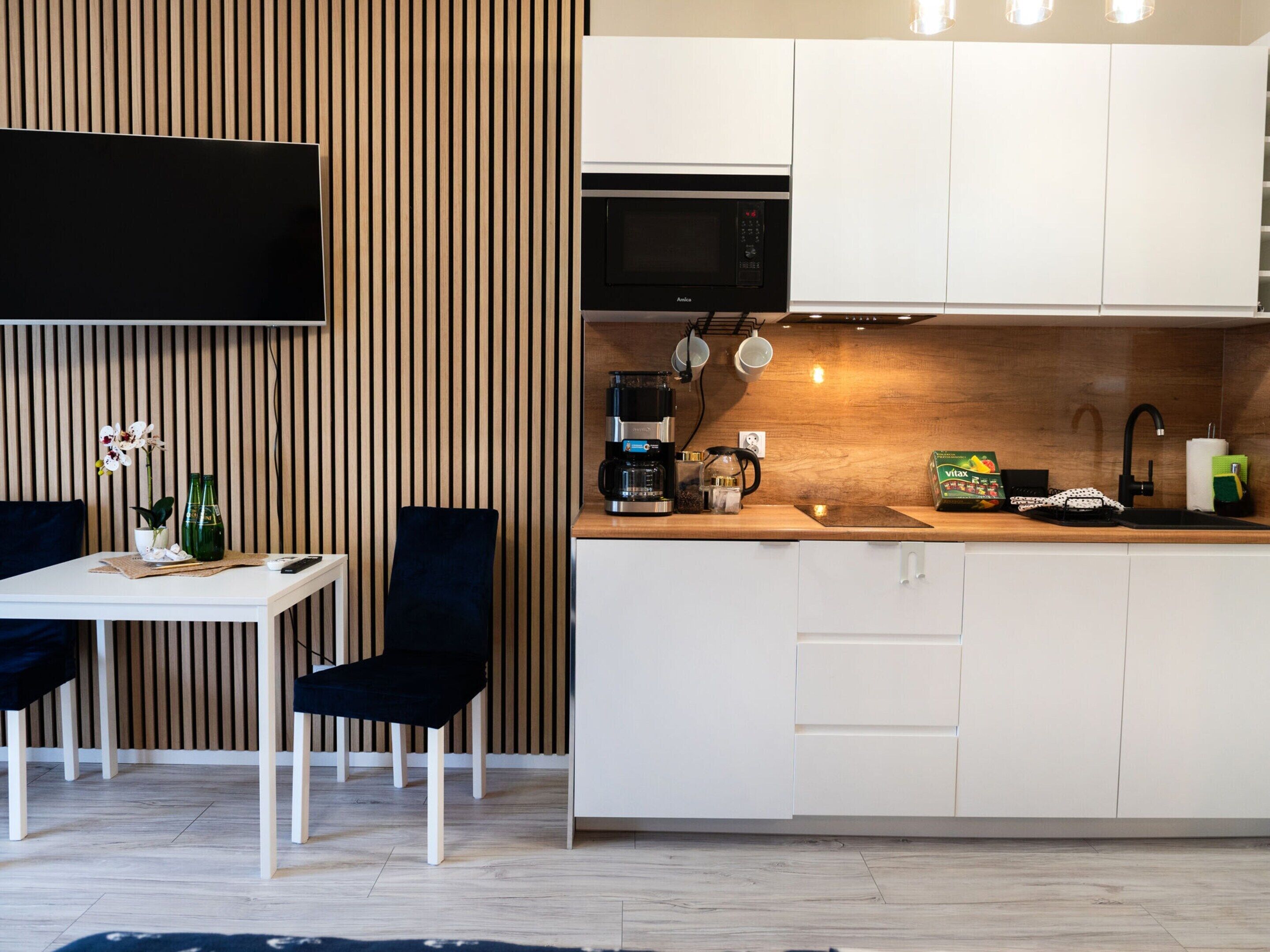 Une cuisine moderne avec des meubles blancs, un micro-ondes noir et une machine à café. Les murs sont recouverts de lambris en bois et il y a un petit coin repas avec une table et des chaises.