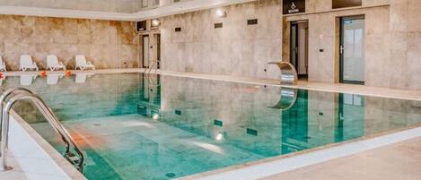 A large indoor swimming pool with a clear water surface, surrounded by a tiled floor and walls. There are lounge chairs along the poolside, and a changing room with a door labeled "Changing Room" is visible.