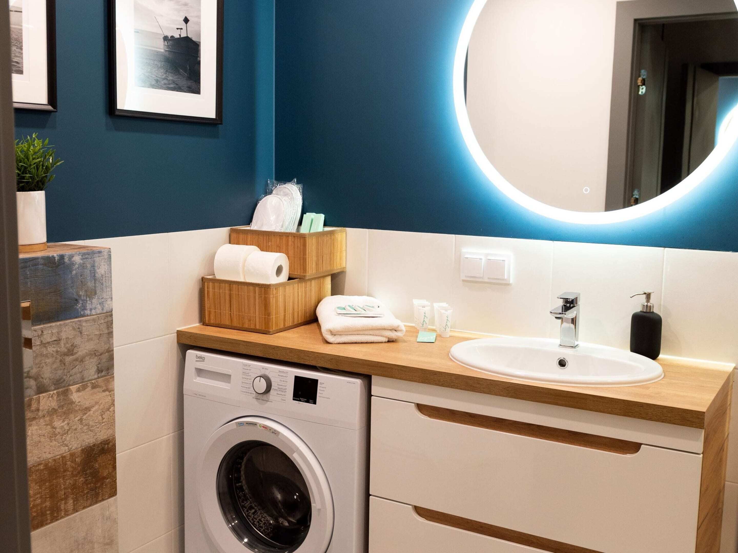 Un baño moderno con lavadora, lavamanos y espejo con luz circular. Hay toallas y artículos de aseo en la encimera.