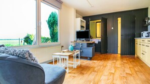 Ein modernes Wohnzimmer mit einem grauen Sofa, einem kleinen weißen Tisch und einem Fernseher auf einem Schrank. Der Raum verfügt über Holzdielen und ein großes Fenster mit Blick auf Grünflächen.
