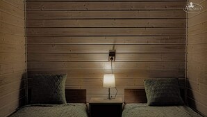 A room with wooden panel walls, two beds with green textured bedding, a nightstand with a lamp, and a wall-mounted electrical outlet. - New Holiday Homes for 2 People in Dziwnowek (Kamien Pomorski)