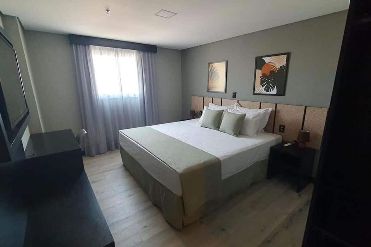 A hotel room with a large bed, a desk, a chair, a TV, and a window with curtains.