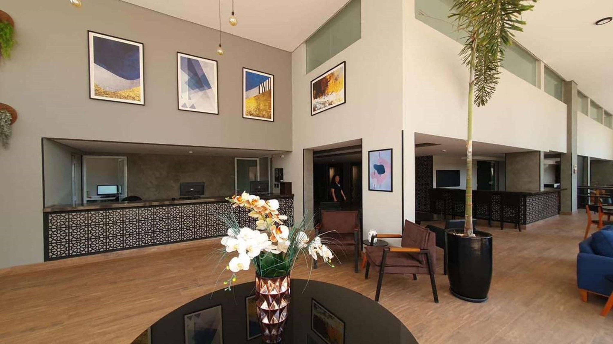 A hotel lobby with a reception desk, framed artwork on the walls, a large potted plant, and a seating area with chairs and a sofa.