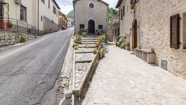 Una calle estrecha que conduce a un edificio de piedra con una torre de campanas, rodeado de muros de piedra y macetas con plantas.