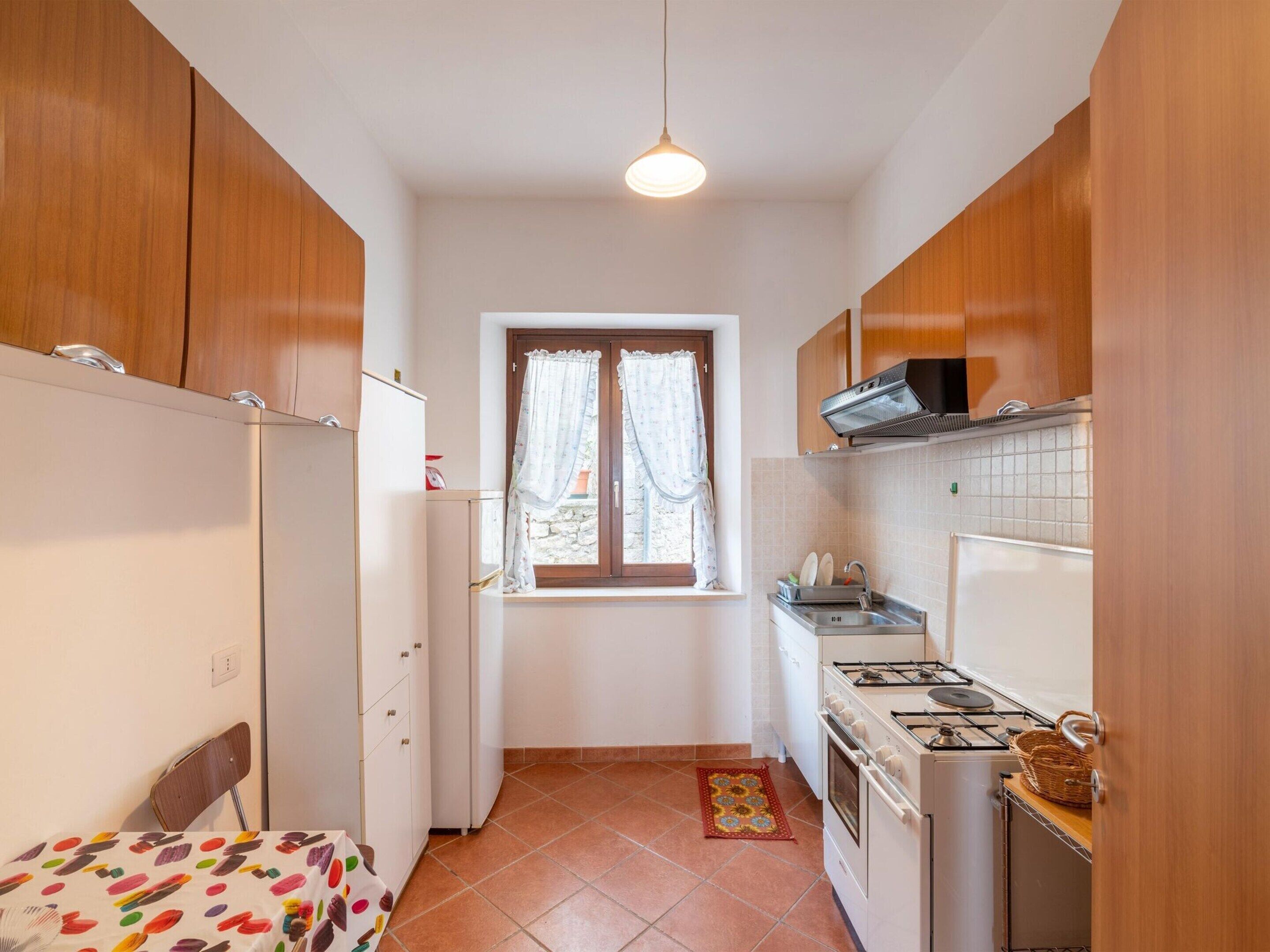 A compact kitchen with wooden cabinets, a white refrigerator, a stove, and a window with curtains.