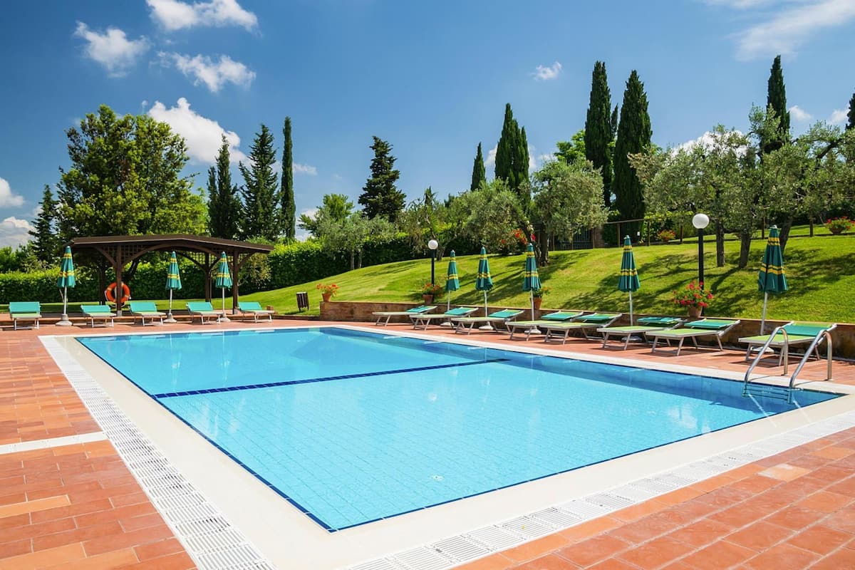 A rectangular swimming pool with a tiled deck, surrounded by green lawn chairs and umbrellas, with trees and a gazebo in the background.
