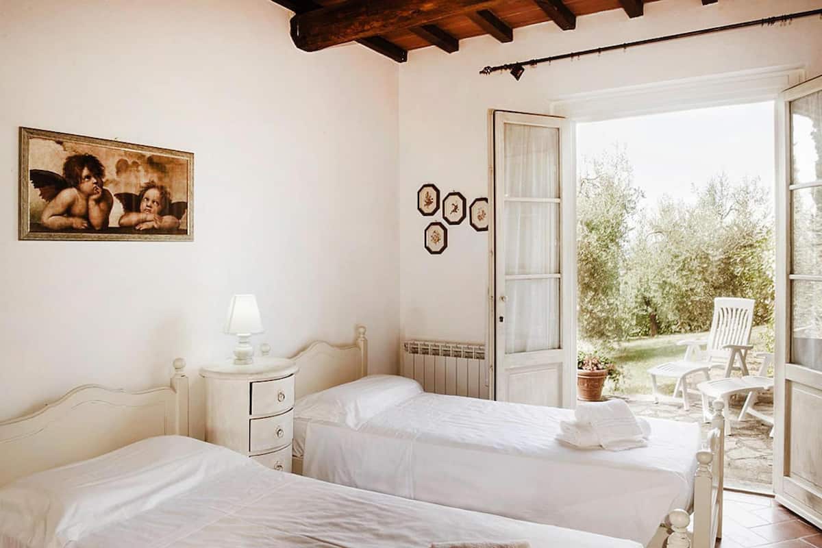 A bedroom with two beds, a painting, a window, and a door leading to an outdoor area with chairs.