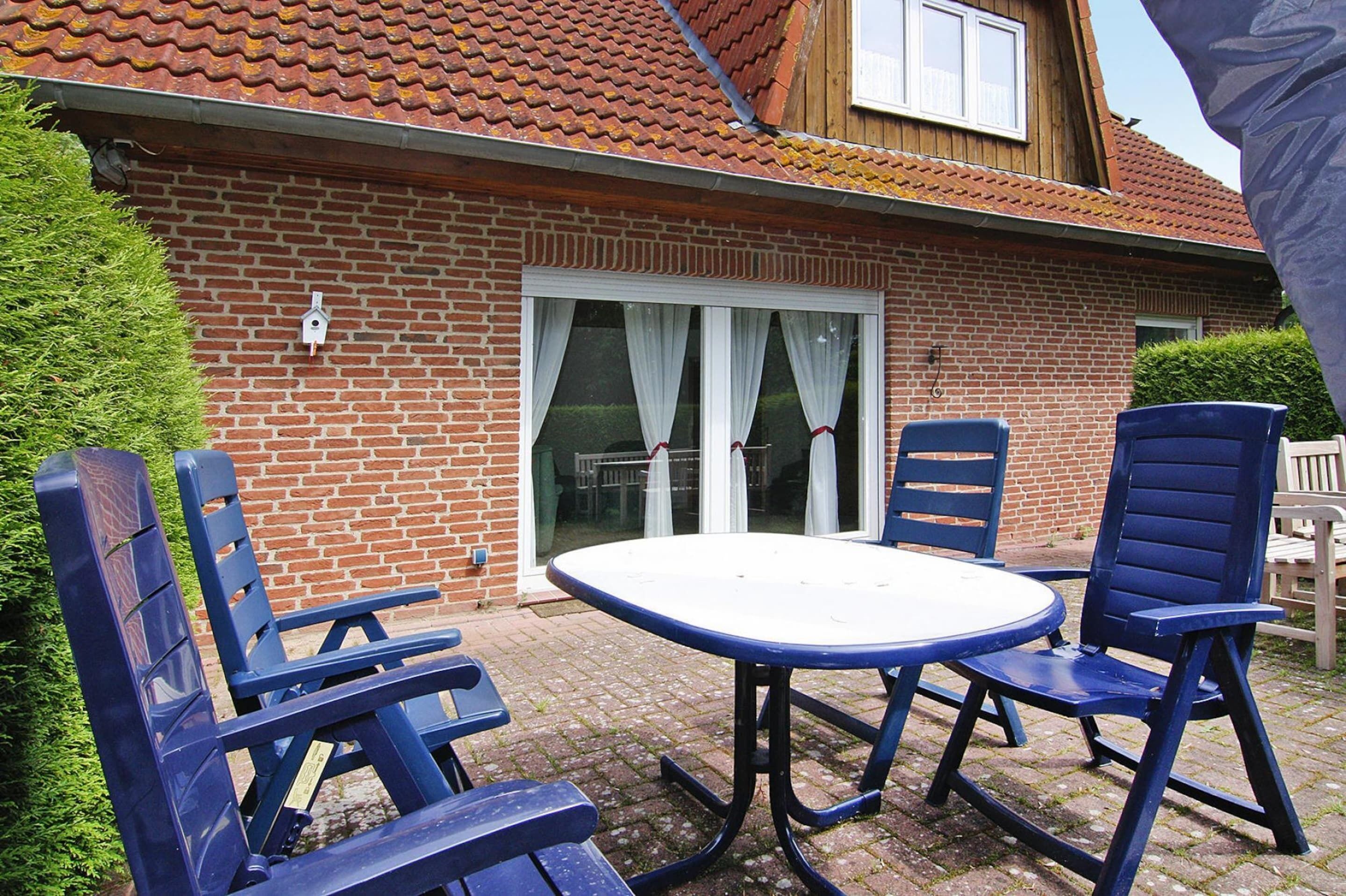 A patio with a round table and four blue chairs, a brick house with a red-tiled roof, and a white window with curtains.