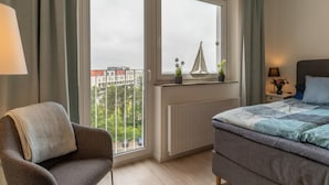 Et soveværelse med en seng, en stol, et vindue der viser udsigt til bygninger, en radiator og et natbord med en lampe.