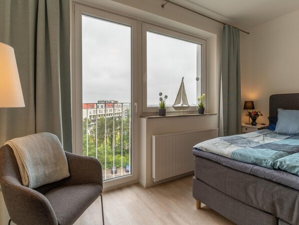 A bedroom with a bed, a chair, a window with a view of buildings, a radiator, and a nightstand with a lamp. - Apartment in St. Peter-ording (St. Peter-Ording)