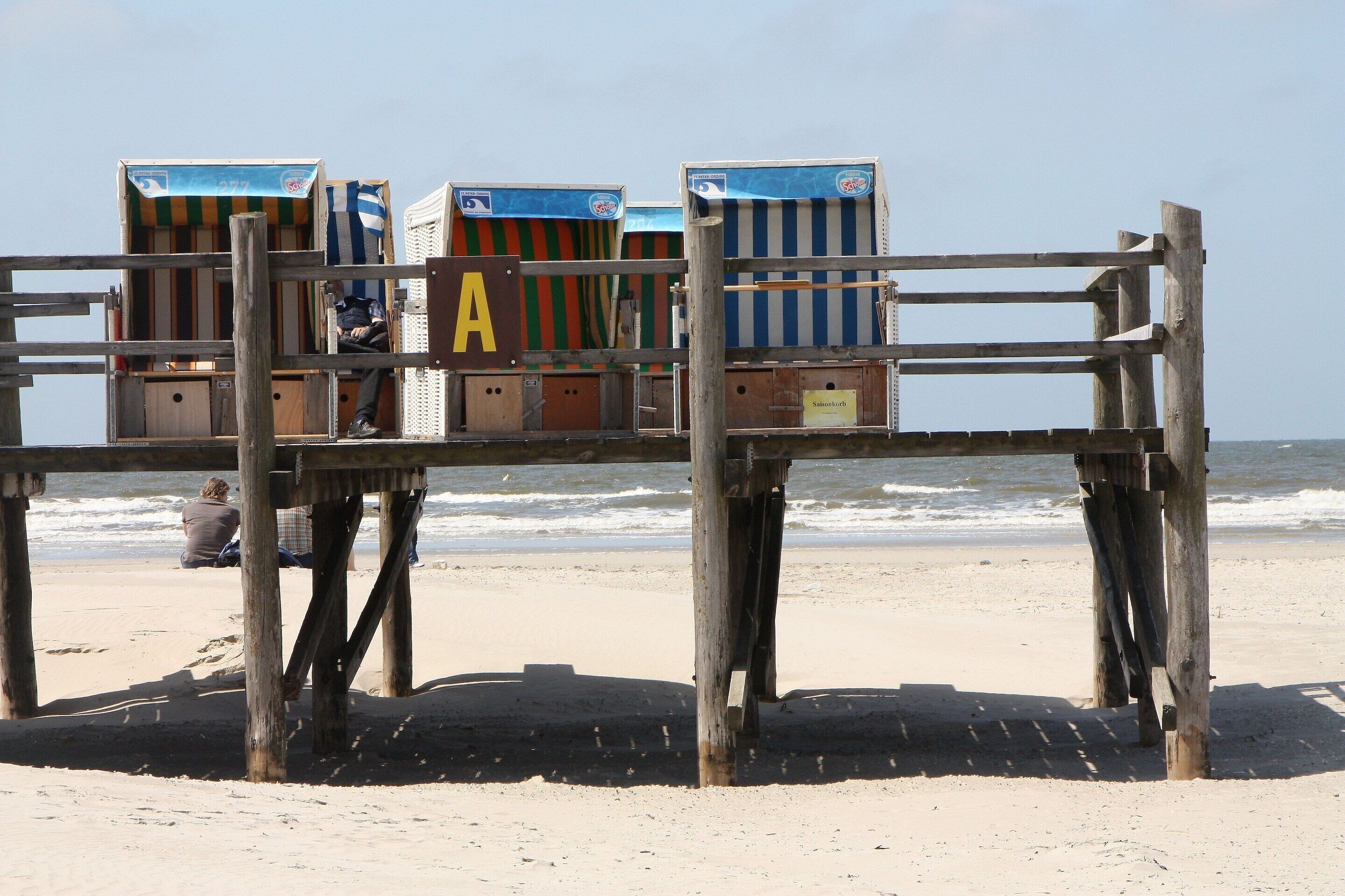 Eine Holzhütte mit Strandbuden, die mit Buchstaben beschriftet sind, ein Sandstrand und das Meer im Hintergrund.
