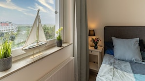 Et soveværelse med en seng, et natbord med en lampe, et vindue med en modelsejlbåd og udsigt til bygninger og træer.