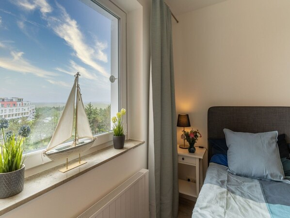 A bedroom with a bed, a nightstand with a lamp, a window with a sailboat model, and a view of buildings and trees. - Apartment in St. Peter-ording (St. Peter-Ording)