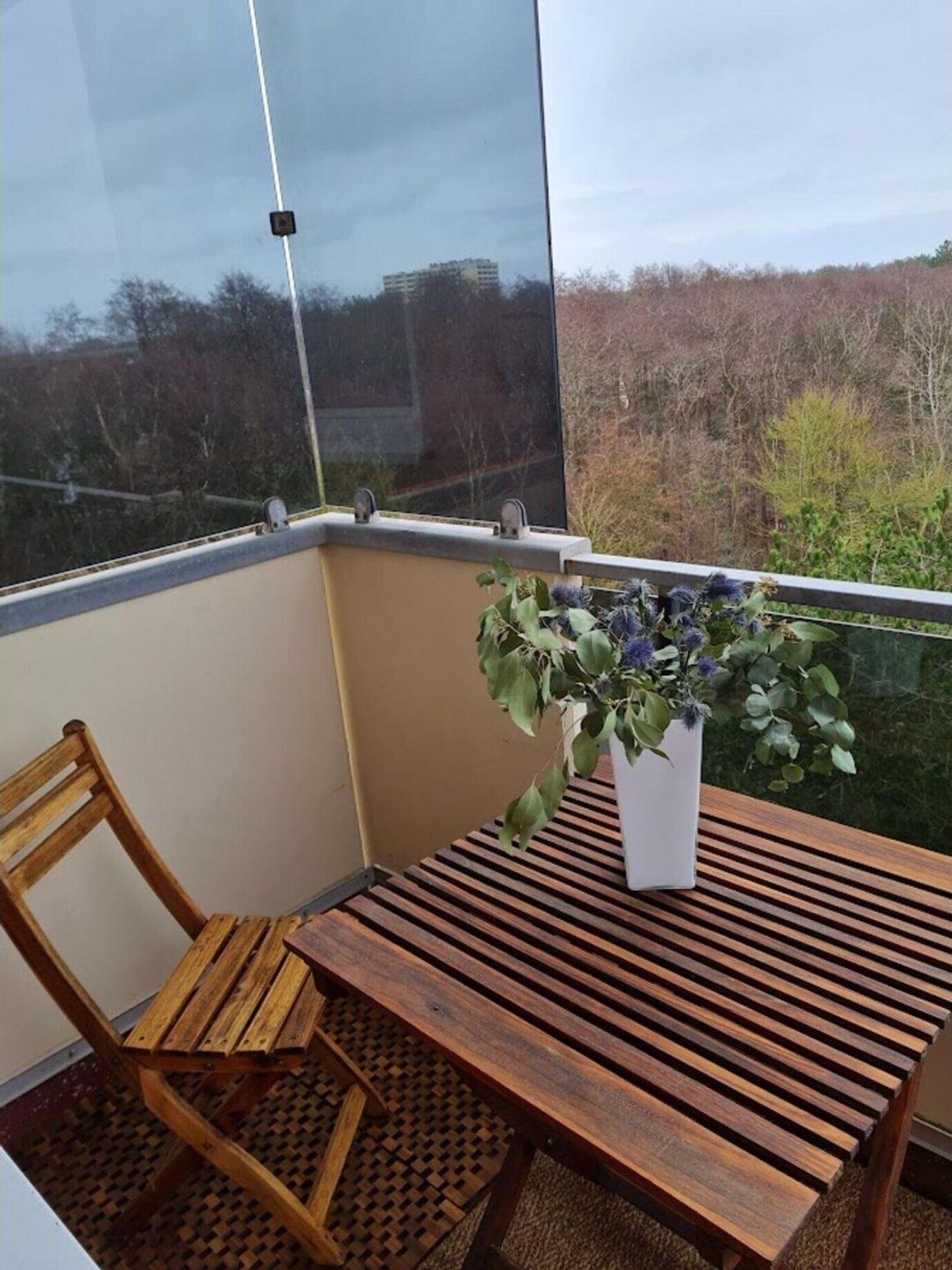 A small balcony with a wooden table and a chair, a potted plant, and a view of trees and buildings.
