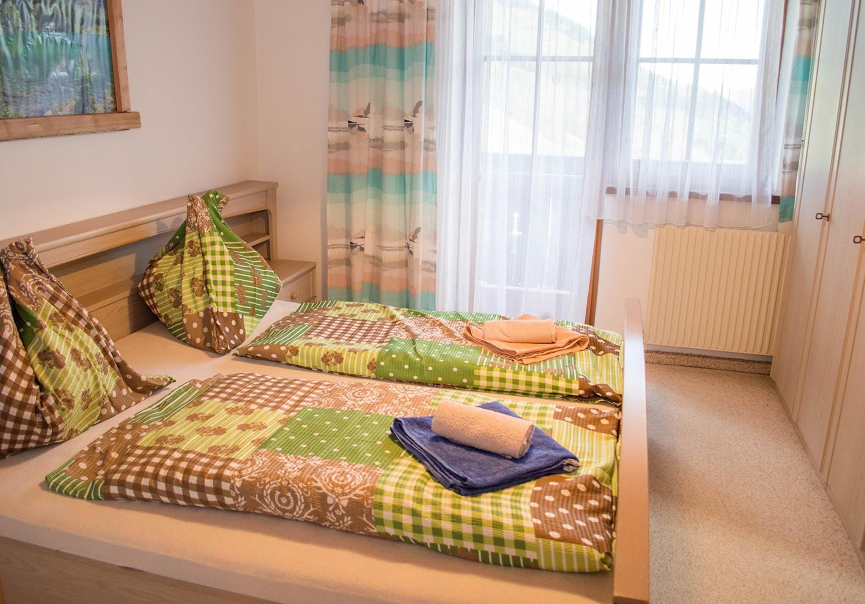 A room with two beds, each with patterned bedding and pillows. There is a window with sheer curtains and a radiator on the wall.