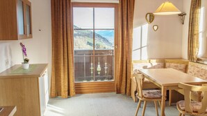 A room with a wooden ceiling, a dining table with chairs, a bench, a small desk, a potted plant, a window with curtains, and a view of a mountainous landscape. - Rauchenbacher Apartments, Mittersill (Mittersill)