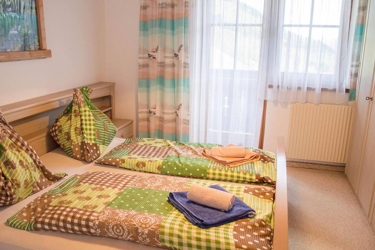 Una habitación pequeña y acogedora con dos camas, cada una con colcha y almohadas a cuadros. Se observa una ventana con cortinas traslúcidas y un radiador en la pared.