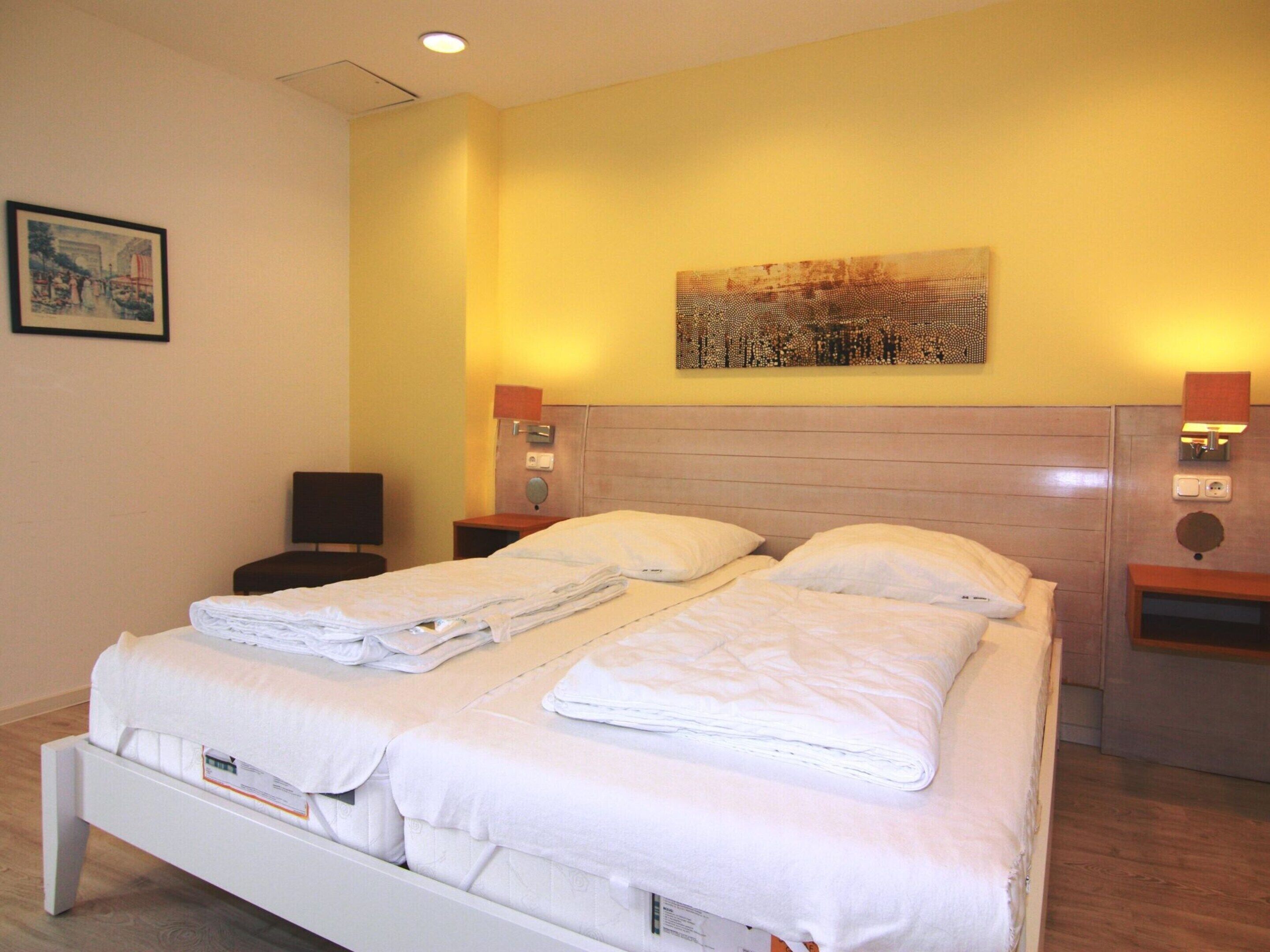 A hotel room with two beds, a headboard, bedside tables, a painting, and a ceiling light.