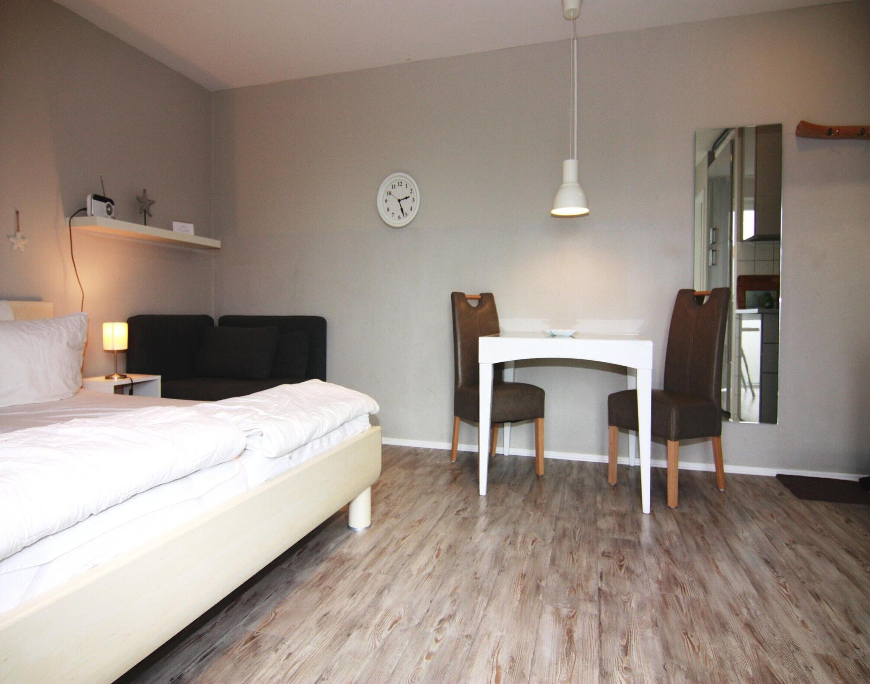 A bedroom with a bed, bedside table, lamp, desk, two chairs, a clock, and a mirror.