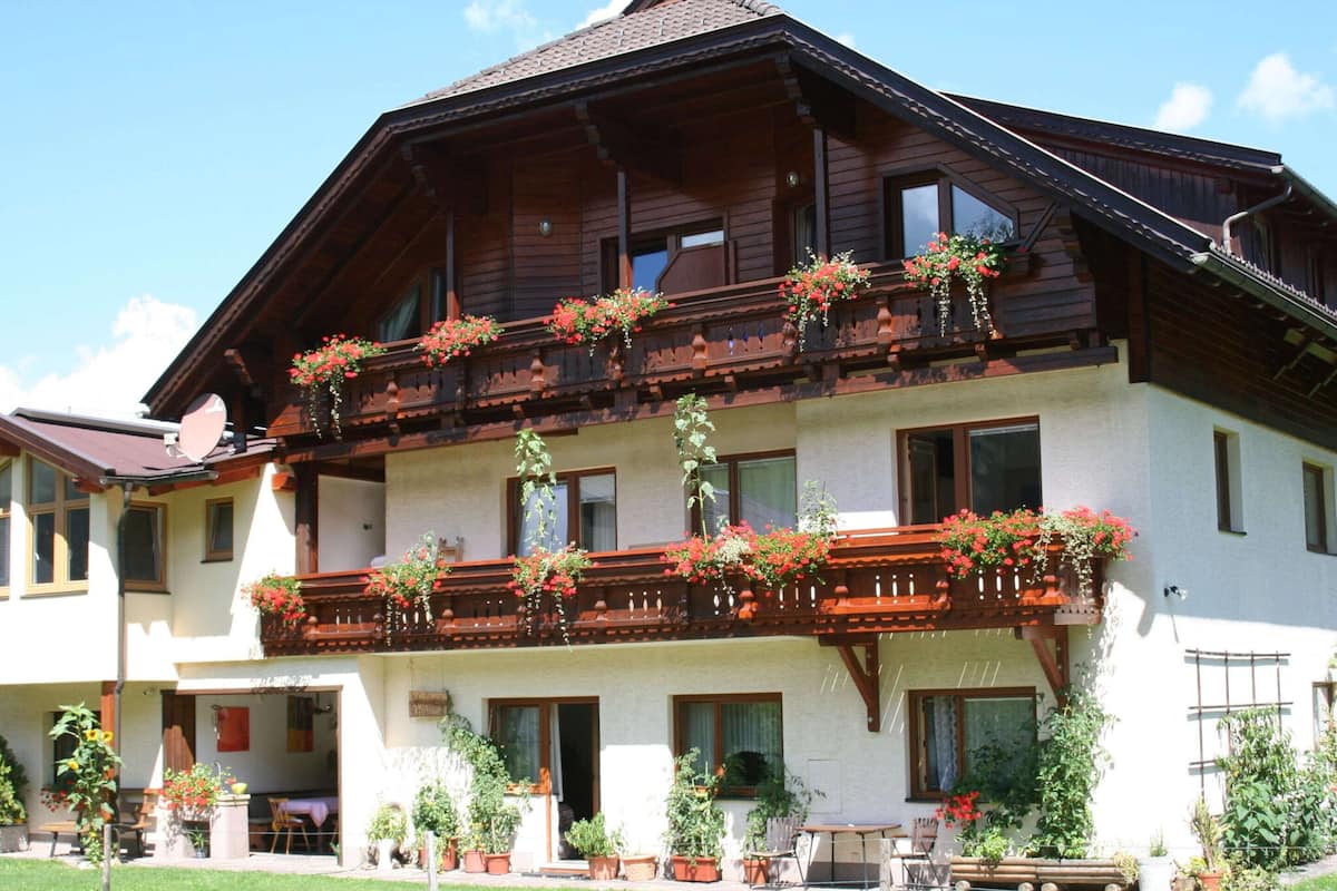 A two-story wooden building with balconies adorned with flowers, a covered outdoor seating area, and potted plants.