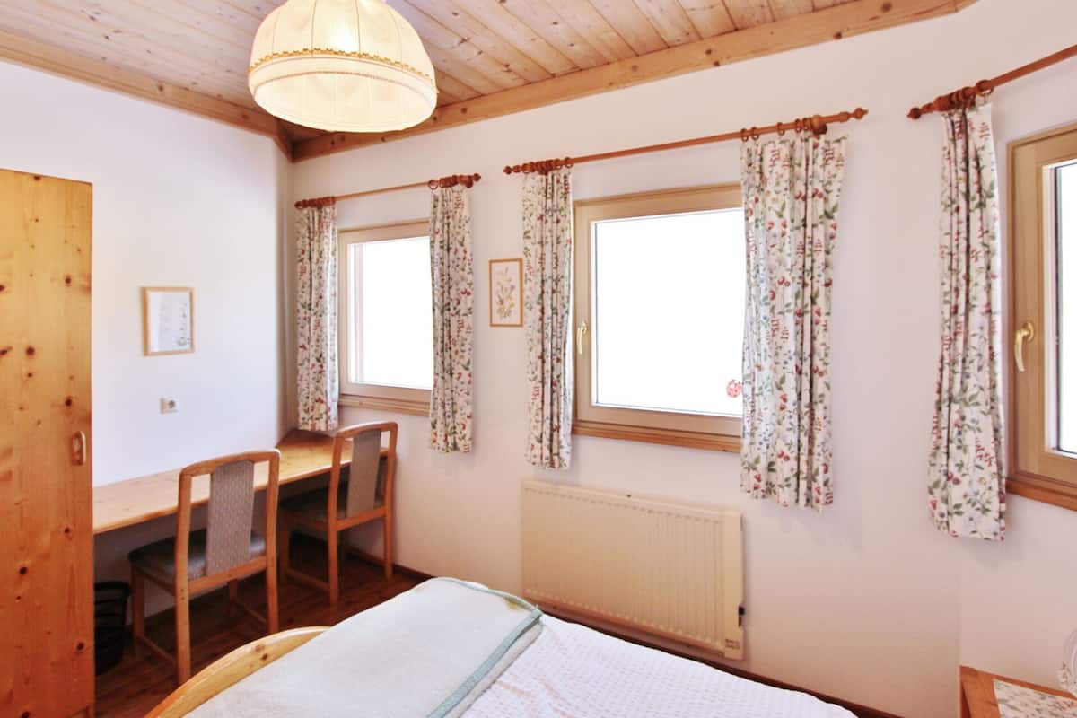 A room with wooden ceiling beams, a bed with a white duvet, a desk with a chair, and two windows with floral curtains.