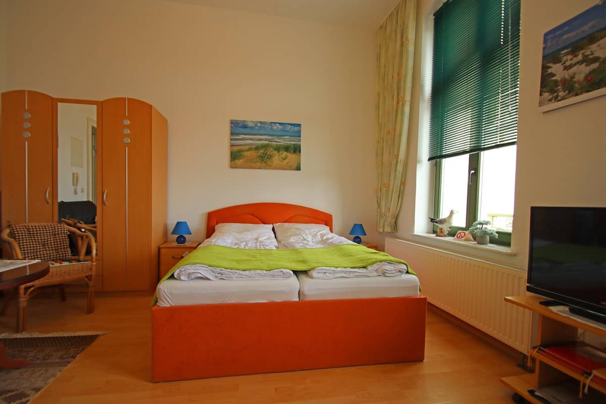 A bedroom with a large bed, a television, a window with blinds, a chair, and a painting on the wall.