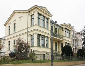 Um edifício grande e cor de creme, com várias janelas e varandas, apresentando elementos arquitetônicos clássicos e fachada simétrica.