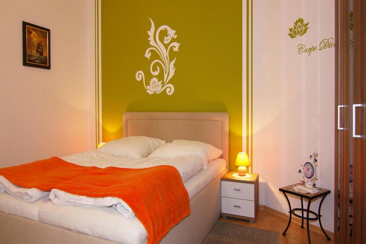 A neatly made bed with an orange blanket, a bedside lamp, and a small table with a clock.