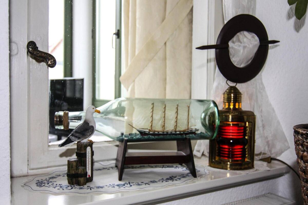 A window sill with a glass ship model, a bird figurine, a decorative plate, and a red candle holder.