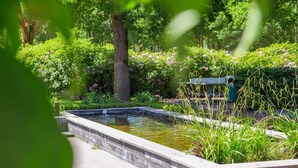Un jardín tranquilo con un estanque rectangular, abundante vegetación y un banco.