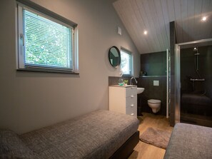A bedroom with a bed, a bedside table, a window with blinds, a mirror, and a bathroom with a toilet and shower. - Cozy Tiny House With a Nice Terrace Near Eindhoven (Oirschot)