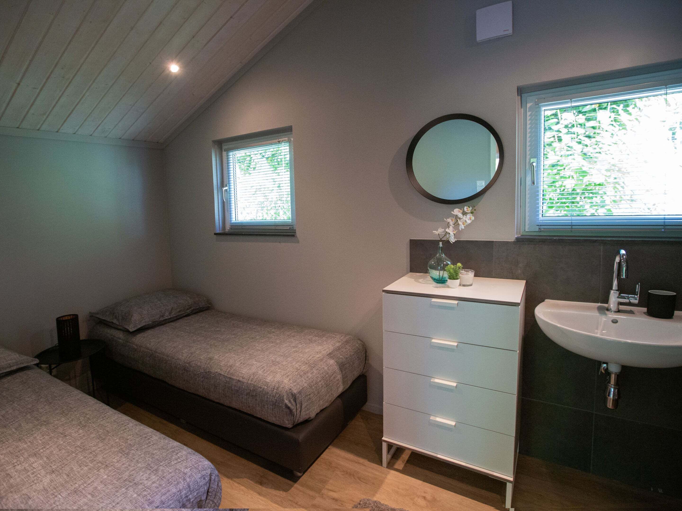 Une chambre à coucher avec un lit, une commode, un miroir rond, une fenêtre et un lavabo.