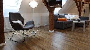A modern loft apartment with exposed wooden beams, a black rocking chair, a grey sofa with orange cushions, a white floor lamp, and a flat-screen TV mounted on the ceiling.