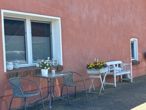 A small outdoor seating area with a table and two chairs, a window with a white frame, and a flower box with yellow flowers. - Charmante Ferienwohnung mit Kamin in Zarchlin (Barkhagen)
