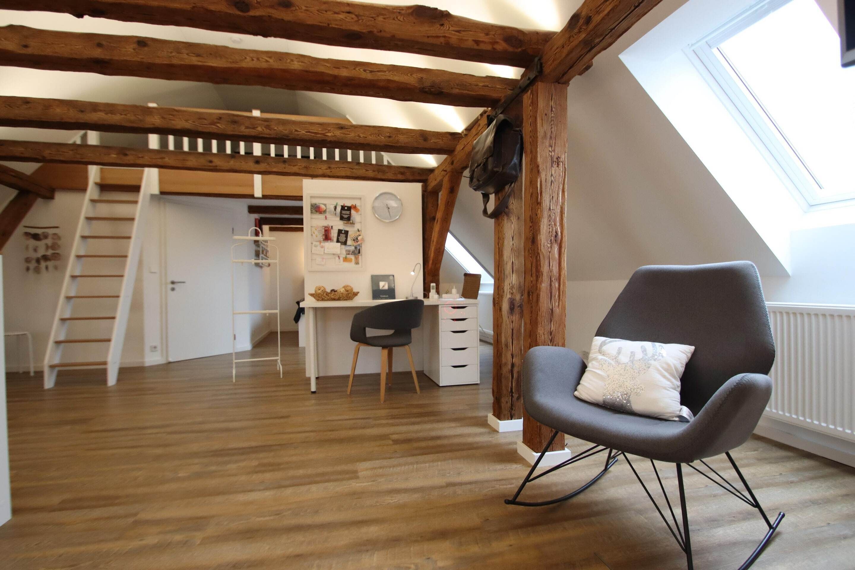 A loft-style room with exposed wooden beams, a desk with a chair, a black rocking chair, and a skylight.