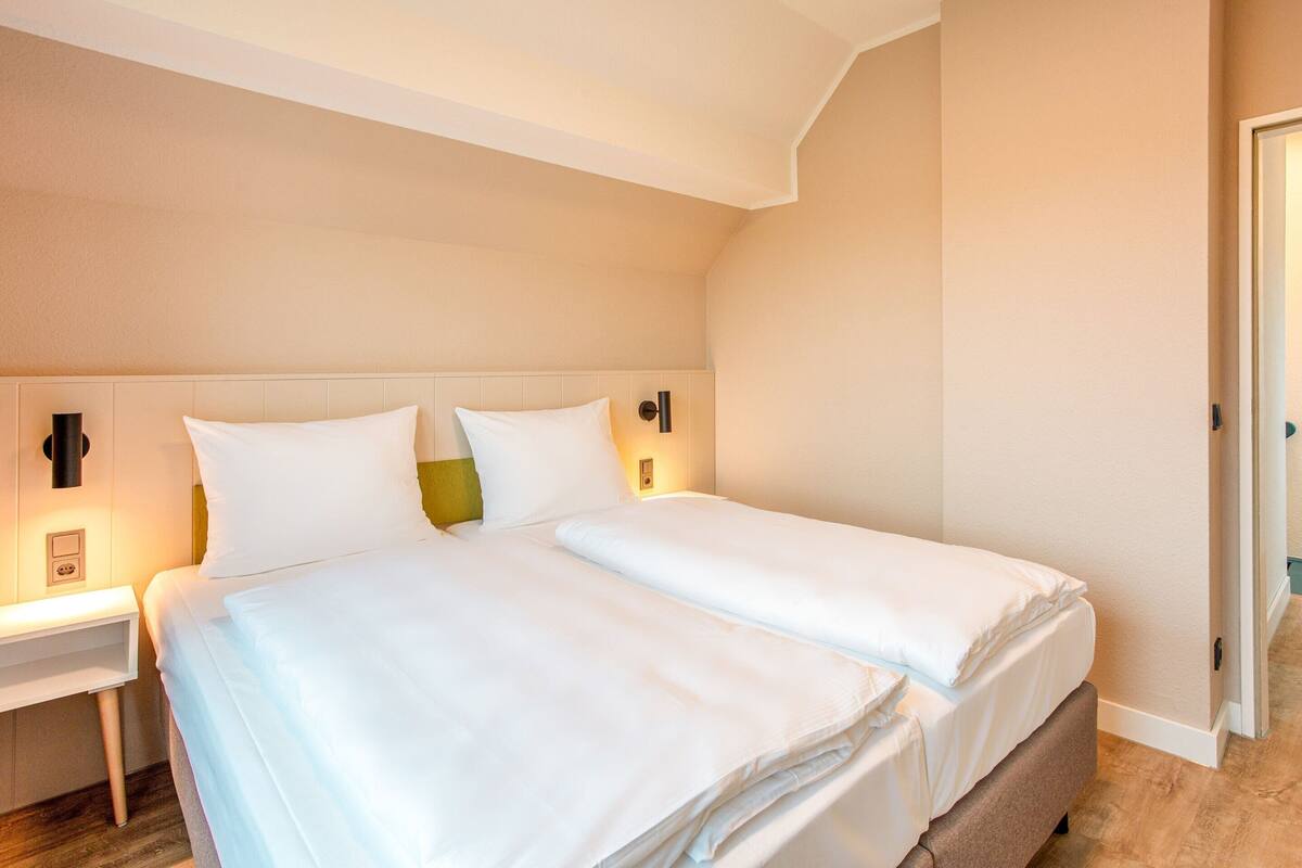 A double bed with white linens, a bedside table with a lamp, and a nightstand with a phone.