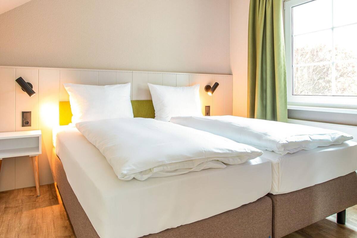 A double bed with white linens, a bedside table with a lamp, and a window with curtains.