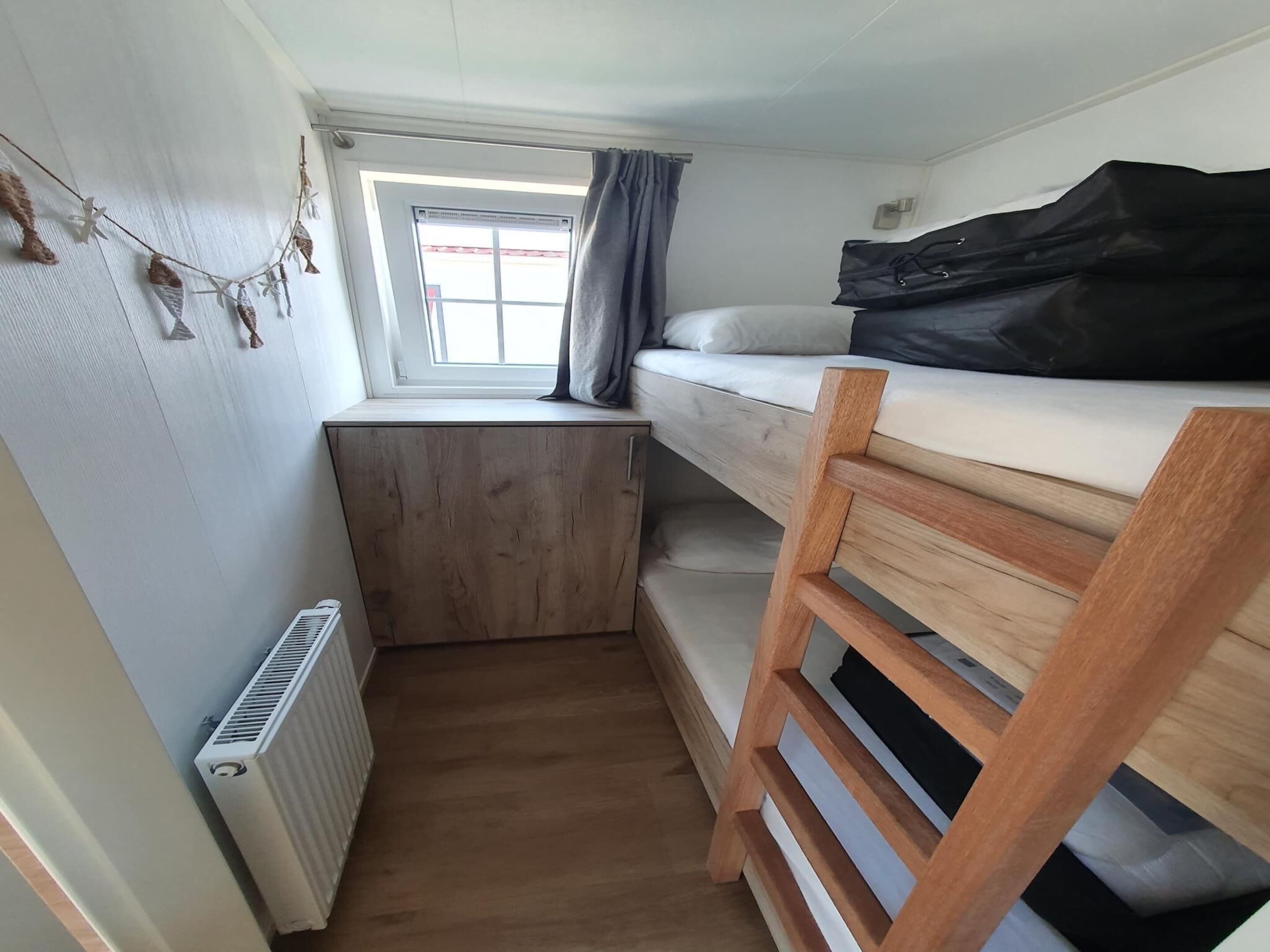 Une pièce compacte et bien éclairée, avec un lit superposé, une petite fenêtre avec des rideaux et un radiateur.