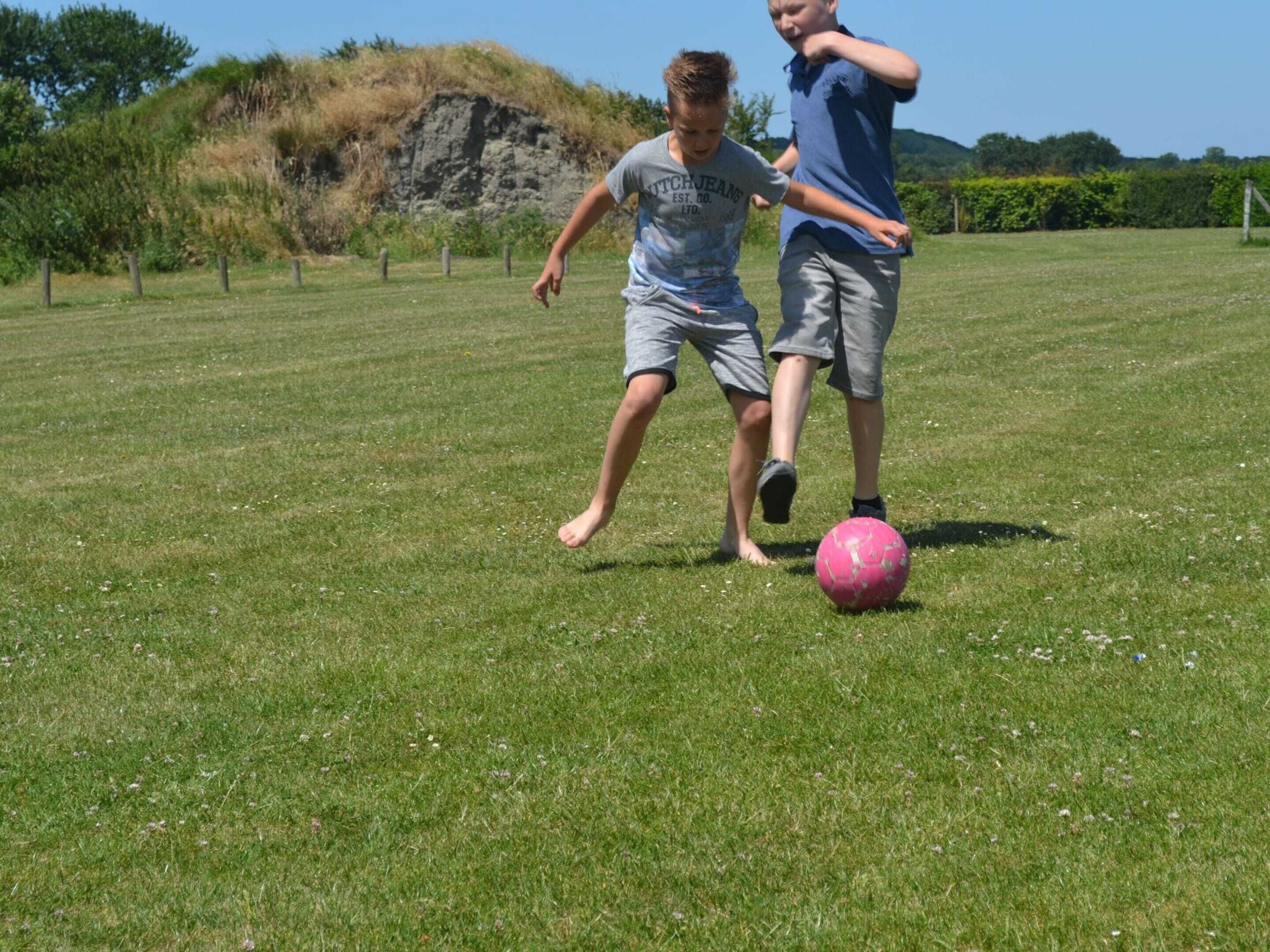 Zwei Kinder spielen Fußball auf einer grünen Wiese mit einem Hügel und Bäumen im Hintergrund.
