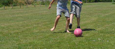 Zwei Kinder spielen Fußball auf einer grünen Wiese mit einem Hügel und Bäumen im Hintergrund.