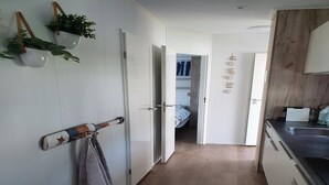 A narrow hallway with a wooden floor, white walls, and a door leading to a bedroom. There are hanging plants and a towel rack on the left wall. - Comfortable Chalet With Airco, on a Holiday Park, at Just 1 km. From the Beach (Koudekerke)