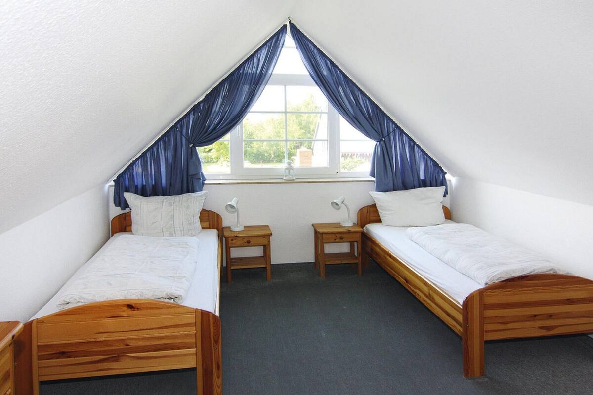 A dormitory room with two wooden beds, a window with blue curtains, and a small table with a lamp between the beds.